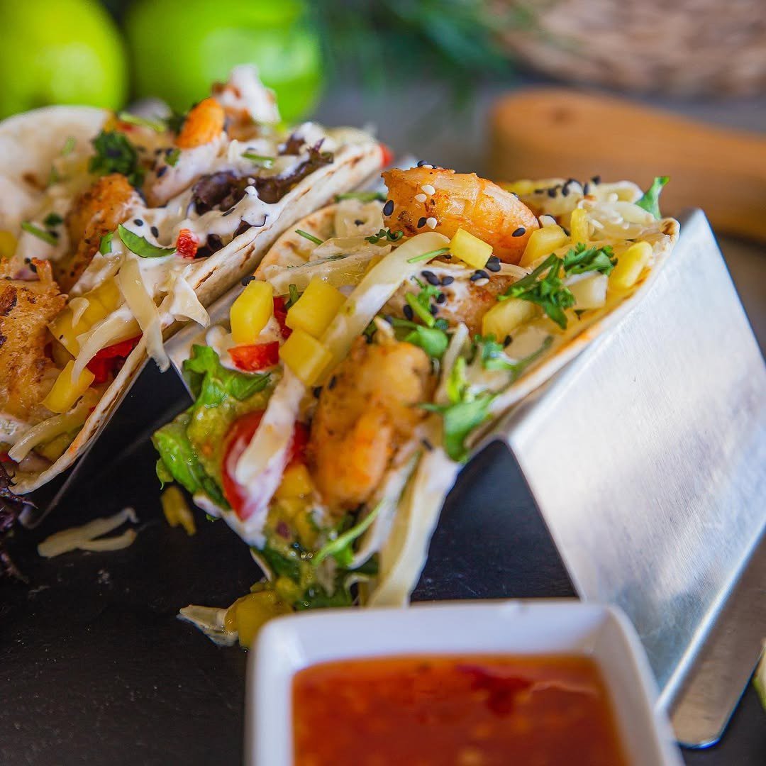 Dois tacos de camarão com pimento e coentros, num suporte de metal, com molho ao lado
