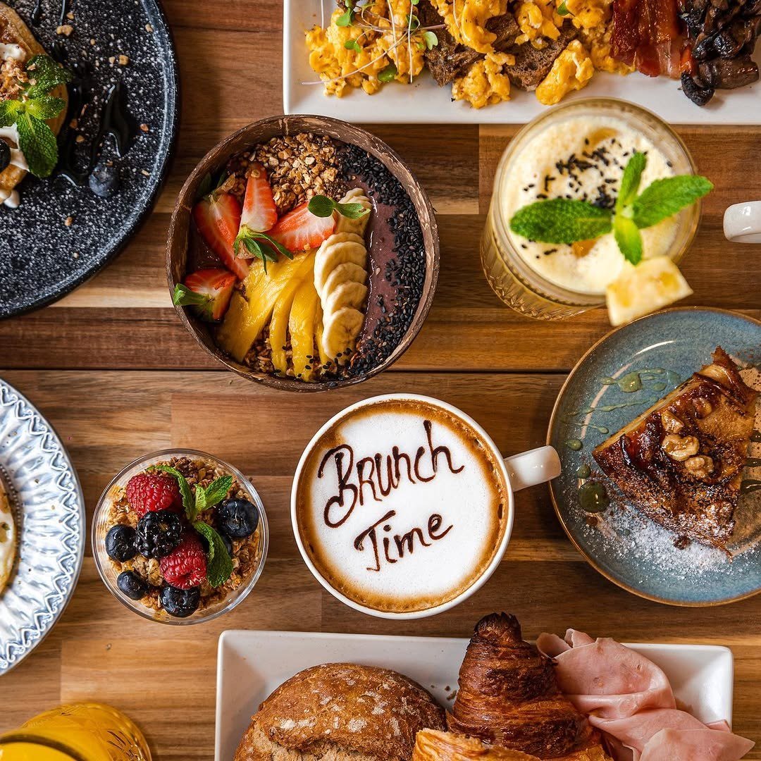 Mesa de brunch vista de cima, com taças de fruta e granola, tostas, croissant e um café com a frase Brunch Time desenhada na espuma